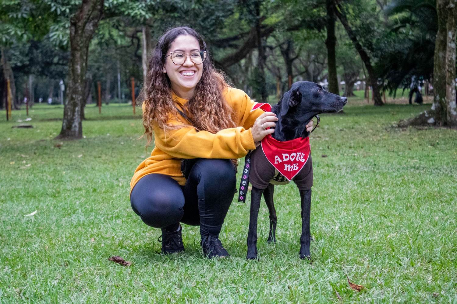 A woman kneeling on the grass beside a black dog wearing an 'Adopt Me' bandana